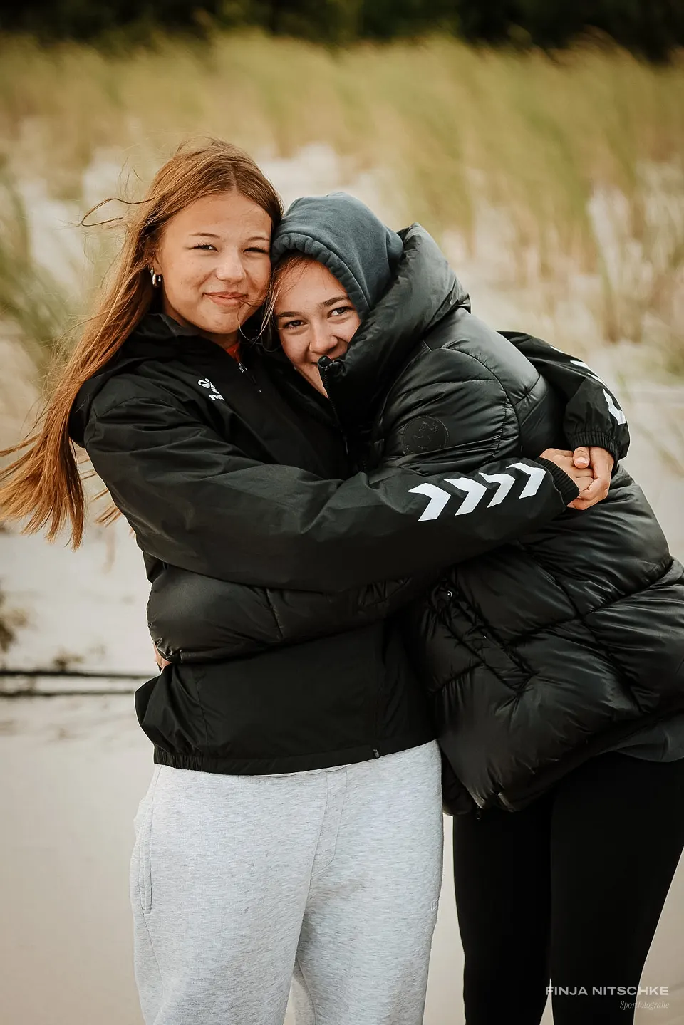 Zwei Sportlerinnen in Winterjacken umarmen sich lachend in den Dünen